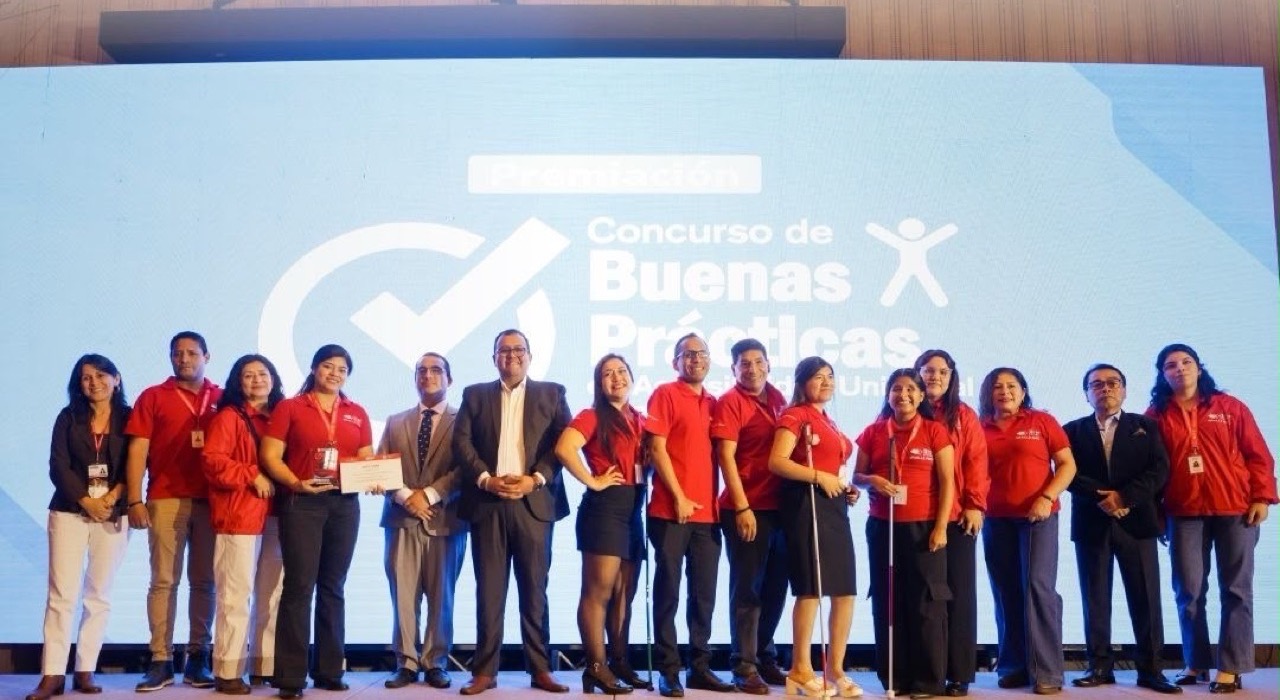 El equipo de Braille Perú sobre el escenario de la ceremonia de premiación, junto a representantes del Ministerio de Vivienda, Construcción y Saneamiento. Quince personas posan en fila; la mayoría lleva el polo rojo de Braille Perú y varias sostienen su bastón blanco. Una integrante sujeta el diploma y el trofeo del primer puesto. Al fondo, la pantalla del Concurso de Buenas Prácticas en Accesibilidad Universal.